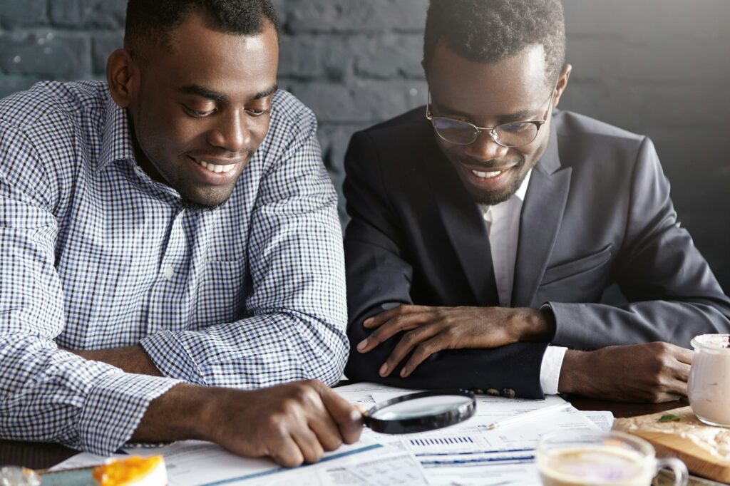 Two happy dark-skinned businessmen reading documents with magnifying glass, analyzing financial agre